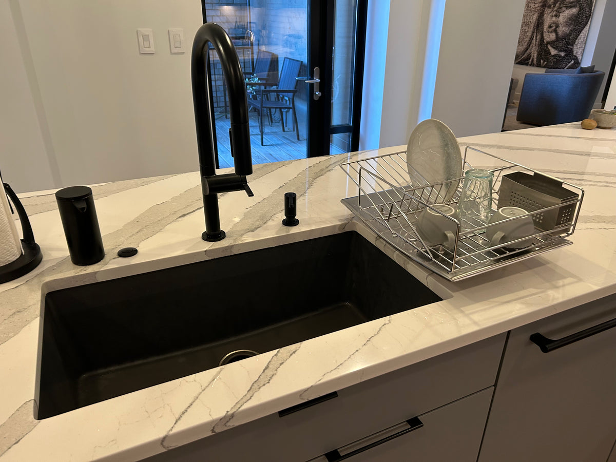 A modern kitchen island featuring a matte black undermount sink, a tall matte black gooseneck faucet, and a stainless steel wire dish rack filled with dishes on a white marble countertop.